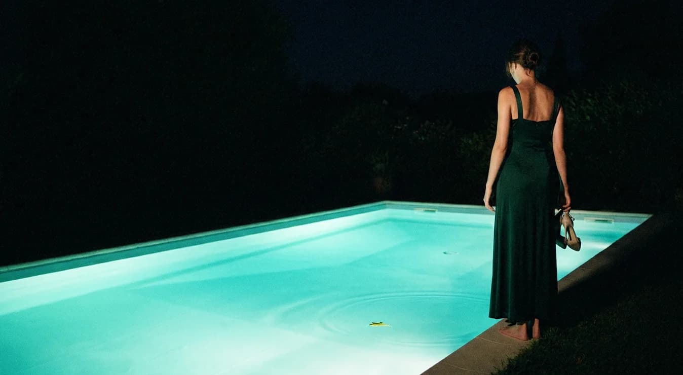 A figure seen from behind at the edge of a glowing pool at night, heels dangling from one hand